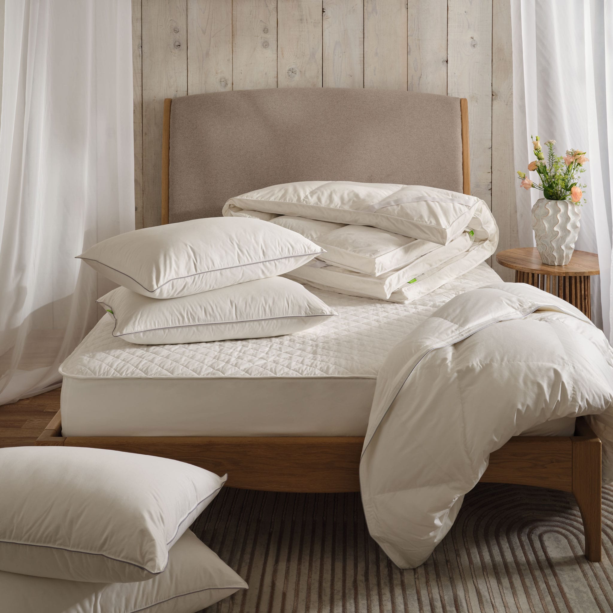 Down pillows, topper and duvet on a bed with a mattress protector. In a light and airy bedroom with net drapes. Scooms