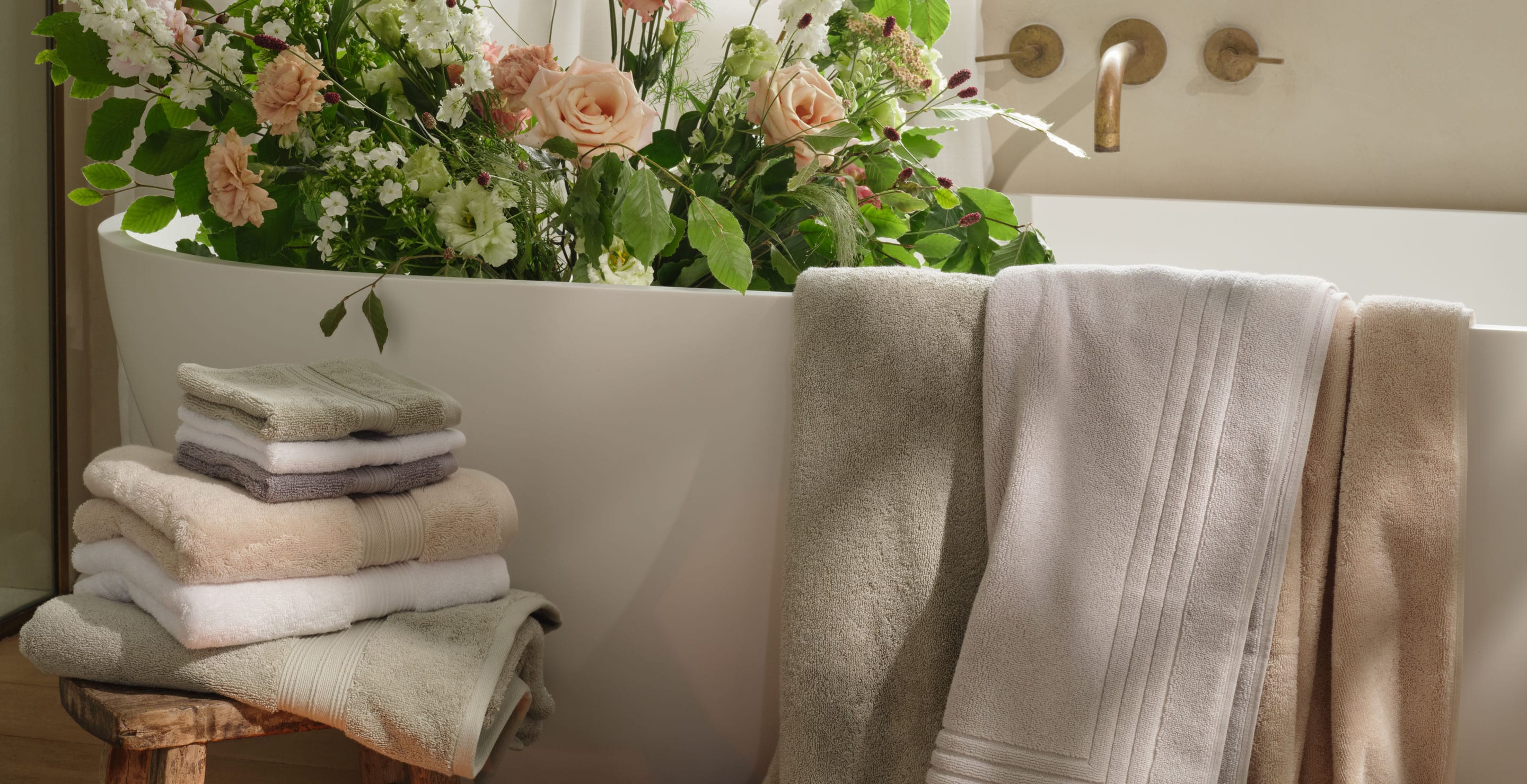 Bathtub with flowers and range of towels in white, green, cream and grey draped over the bath and piled on a stool. | Scooms