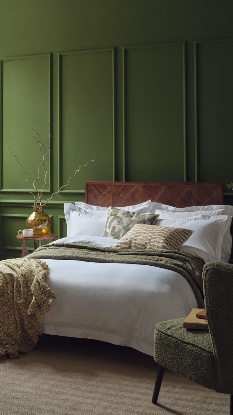 A styled bedroom scene featuring a bed dressed in white Egyptian cotton sateen bedding with patterned accent cushions, set against a dark green panelled wall. A textured green armchair sits in the foreground, and a small side table with a gold vase is visible on the left. | Scooms
