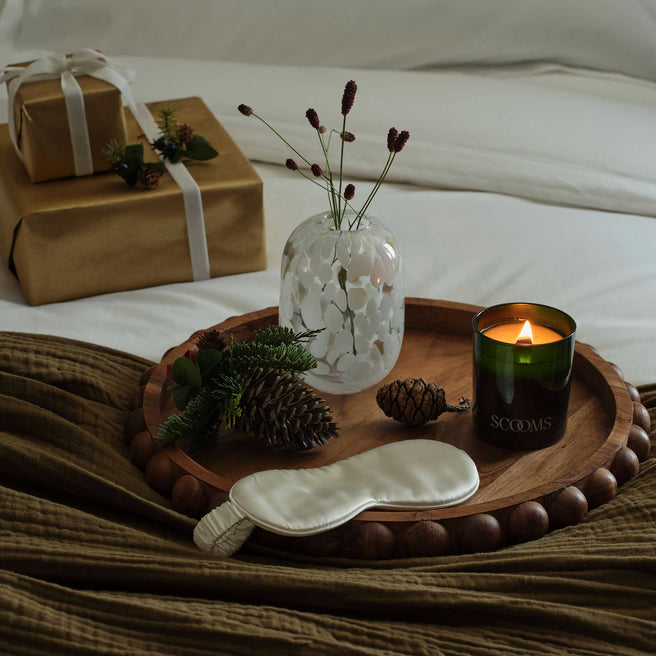 Natural scented candle lit, silk eye mask and decorations on a moss coloured throw on a bed with gift wrapped boxes | Scooms
