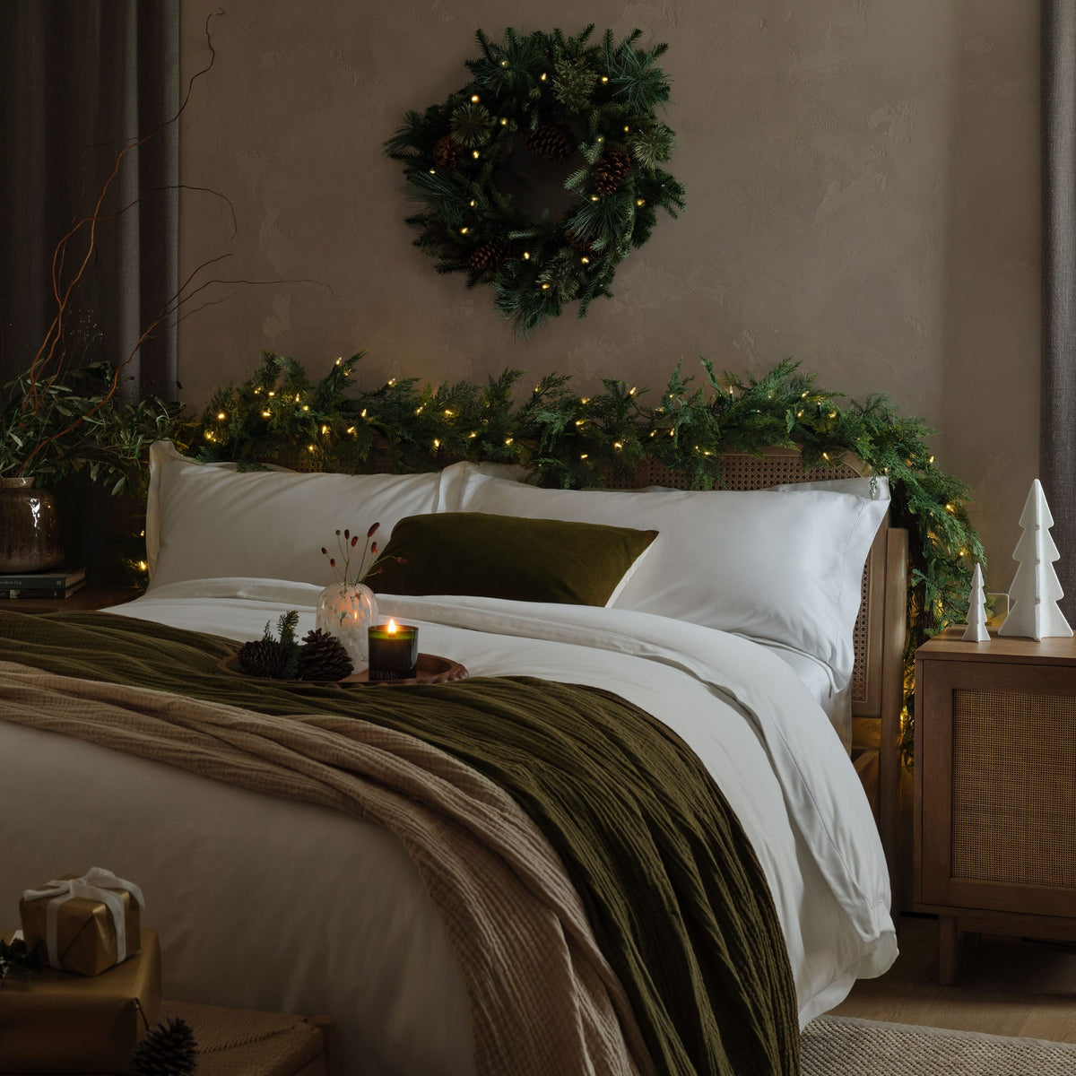 A styled Christmas bedroom scene featuring a bed dressed in white linens with an olive green textured throw blanket and matching pillow, candles, decorated with a green xmas wreath and warm string lights on the headboard, and a wooden nightstand beside it.