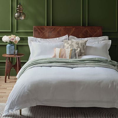 Bedroom with a wooden bed frame, white Egyptian cotton sateen weave bedding, and decorative pillows against a green panelled wall. | Scooms