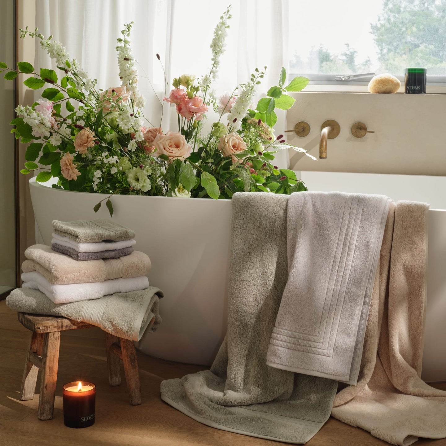 A bathtub with a large arrangement of fresh flowers, folded towels in grey, green, natural and white, stacked on a small wooden stool, and several bath towels in sage green, white and natural colours draped over the tub edge. | Scooms