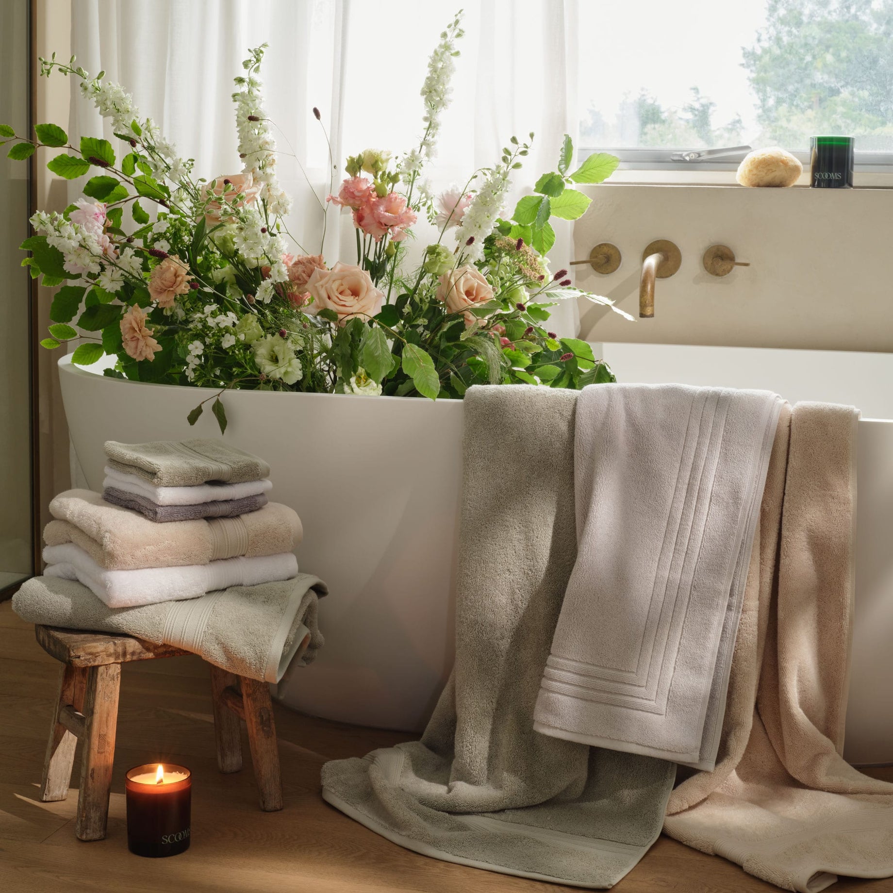 A bathtub with a large arrangement of fresh flowers, folded towels in grey, green, natural and white, stacked on a small wooden stool, and several bath towels in sage green, white and natural colours draped over the tub edge. | Scooms