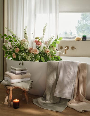 A freestanding bathtub filled with a large floral arrangement. White, natural, green, cream and grey towels are draped over the tub and stacked on a small wooden stool beside it. | Scooms