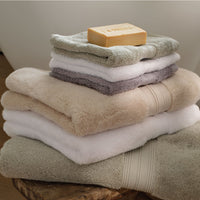 A stack of folded bath towels in neutral tones including natural, white, grey and sage green, with a bar of soap resting on the top towel, set on a wooden surface against a soft neutral background. | Scooms