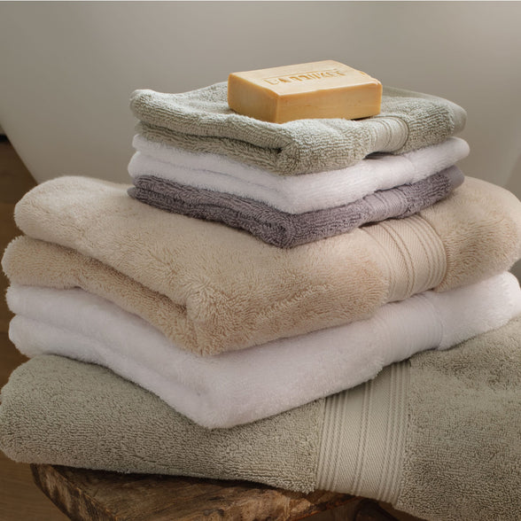 A stack of folded bath towels in neutral tones including natural, white, grey and sage green, with a bar of soap resting on the top towel, set on a wooden surface against a soft neutral background. | Scooms