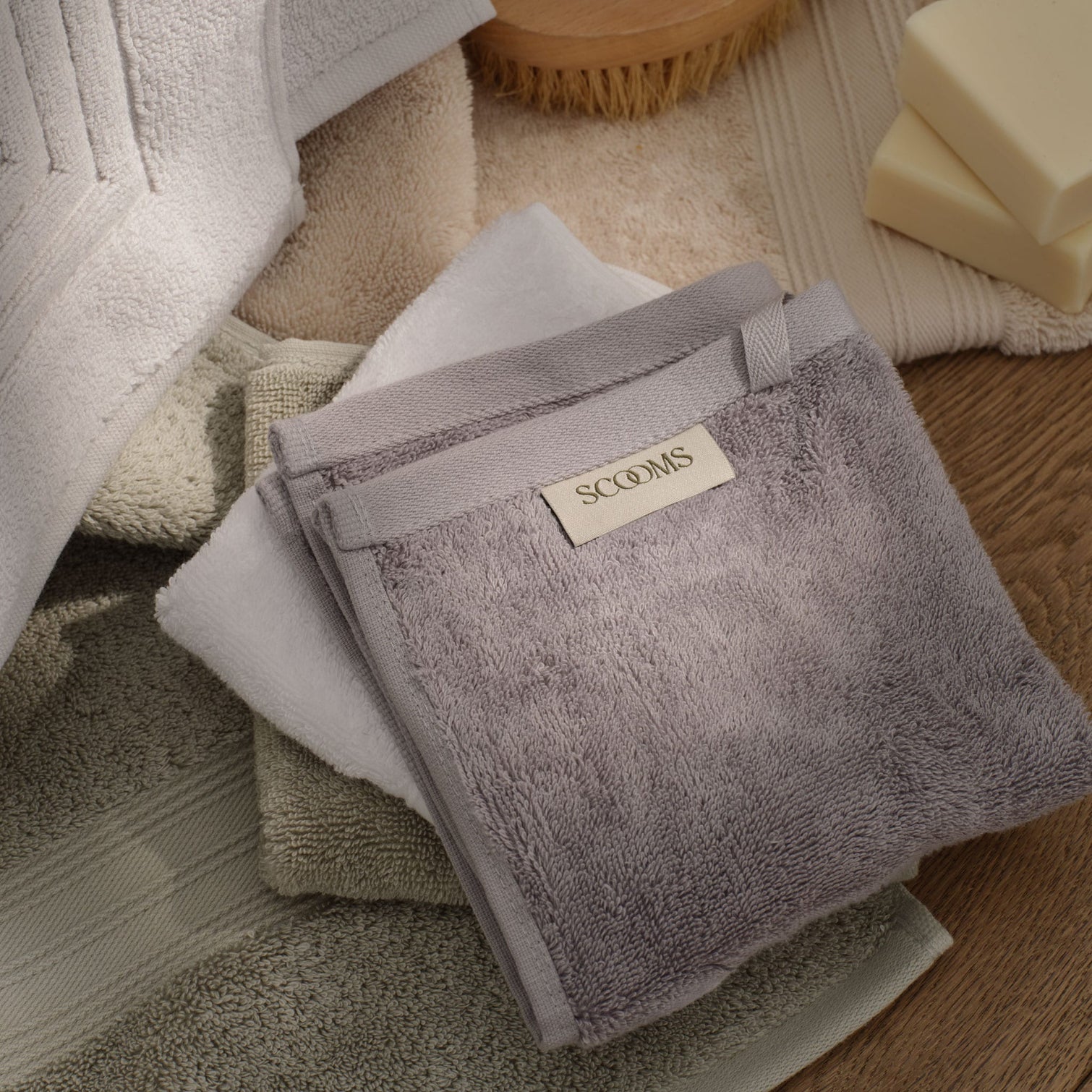 A stack of folded bath towels in soft neutral tones, with a grey towel on top featuring a 'SCOOMS' label. The towels are arranged on a wooden surface with a soap dish and wooden brush visible in the background. | Scooms
