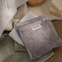 A stack of folded bath towels in soft neutral tones, with a grey towel on top featuring a 'SCOOMS' label. The towels are arranged on a wooden surface with a soap dish and wooden brush visible in the background. | Scooms