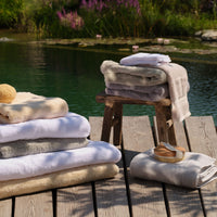 A stack of folded bath towels in white, cream, green, natural and grey tones arranged on a wooden dock beside a calm lake, with a rustic stool holding more folded towels and a small soap dish nearby. | Scooms