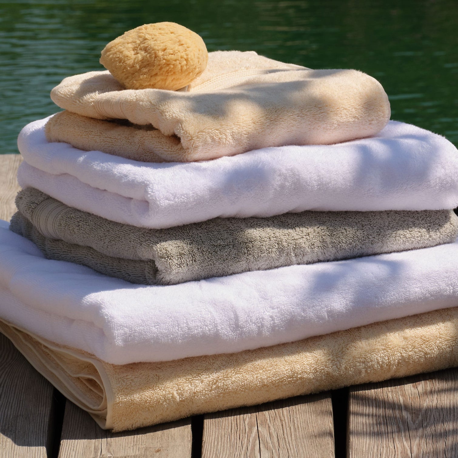 A stack of folded cotton towels in white, light grey, natural and cream, with a natural sponge resting on top, set on a wooden deck with green water in the background. | Scooms