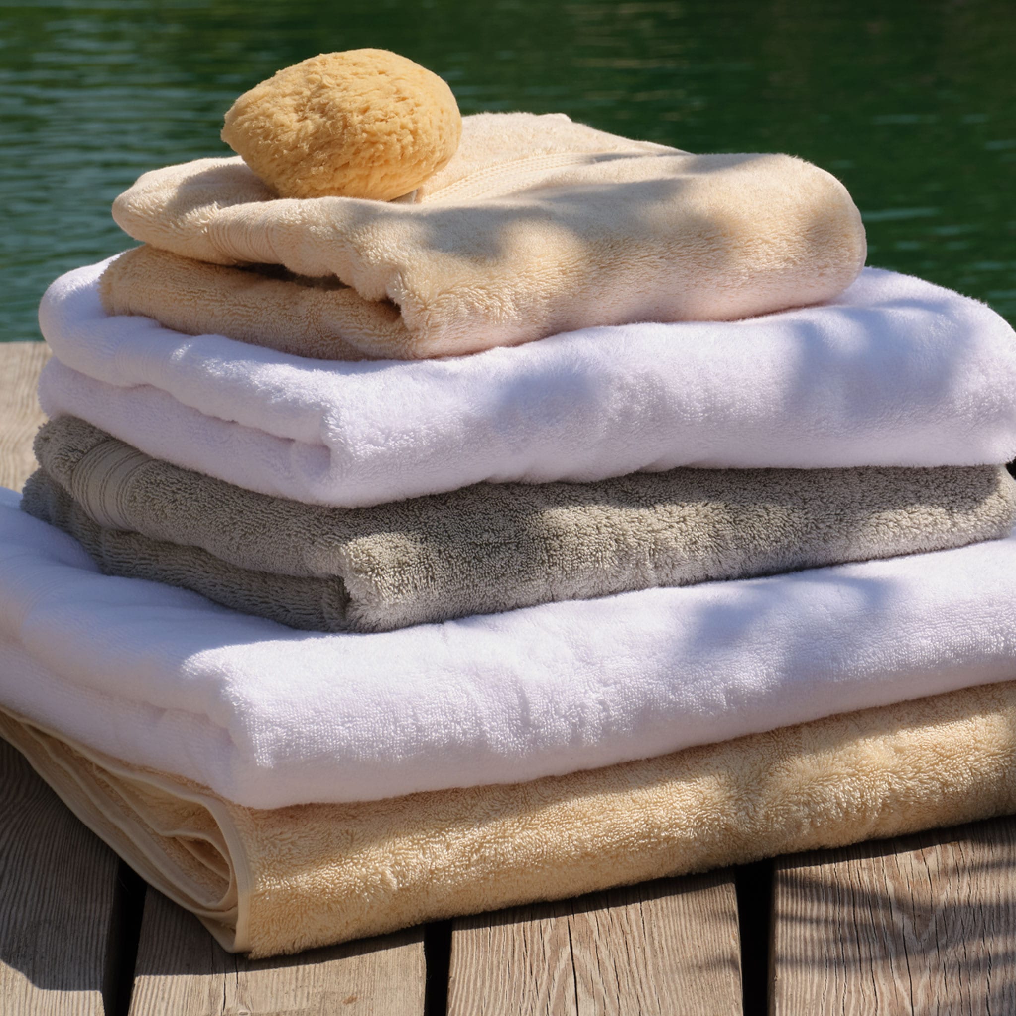 A stack of folded cotton towels in white, light grey, natural and cream, with a natural sponge resting on top, set on a wooden deck with green water in the background. | Scooms