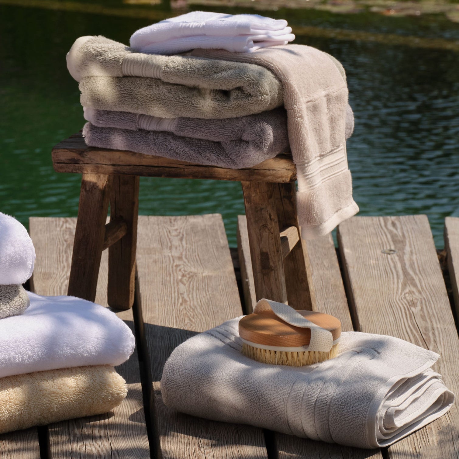 A stack of folded bath towels in white, cream, grey and green tones is arranged on a rustic wooden stool and tabletop beside a body of water. A wooden-handled bath sponge rests on a light grey bath mat in the foreground. | Scooms