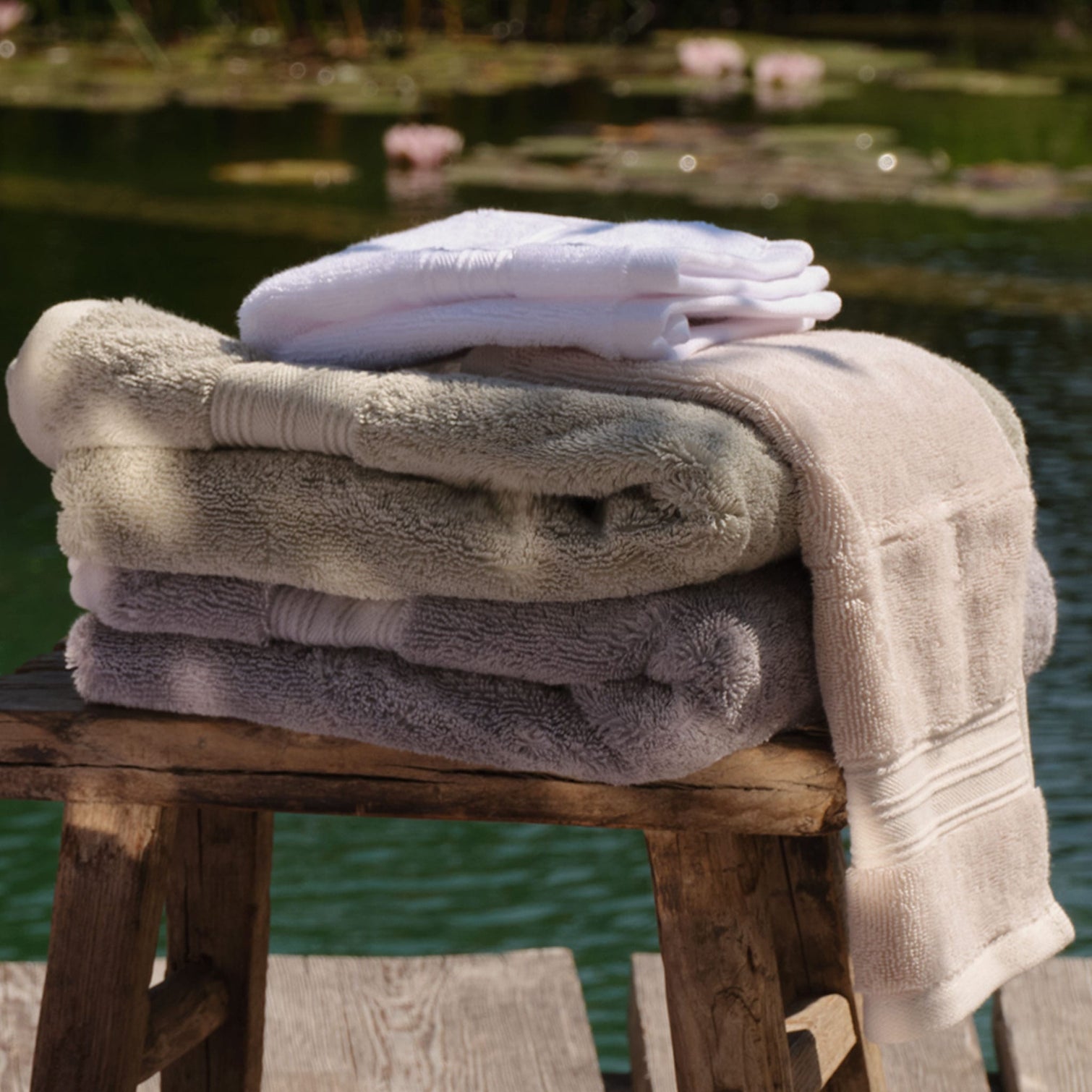 Three folded stacks of plush bath towels in white, light grey, green, cream and natural, arranged on a rustic wooden stool with a pond and lily pads in the background. | Scooms