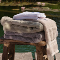 Three folded stacks of plush bath towels in white, light grey, green, cream and natural, arranged on a rustic wooden stool with a pond and lily pads in the background. | Scooms