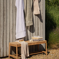 A wooden bench outdoors with folded and hanging towels in neutral tones of white, green, cream, natural and grey. A small soap dish with a brush and folded towels sit on the bench, with greenery and gravel visible in the background. | Scooms