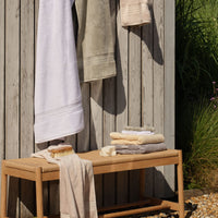 A wooden bench with white, green, natural, cream and white hanging towels, folded towels, and a bath brush arranged on and around the bench in a sunlit outdoor setting. | Scooms