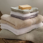 A stack of folded bath towels and face cloths in neutral tones, including sage green, white, natural and grey, with a bar of soap resting on top, displayed on a wooden stool against a soft neutral background. | Scooms