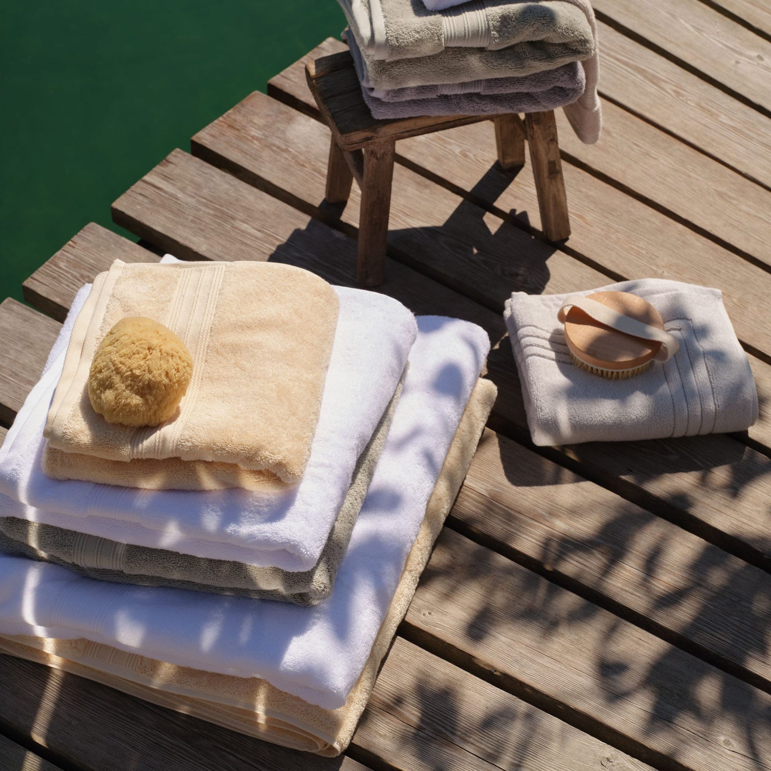 A set of folded Egyptian cotton bath towels and bath mats in cream, white, green and light grey, arranged on a wooden dock next to a green body of water. One stack includes a beige loofah sponge and another includes a wooden-handled towel brush. | Scooms