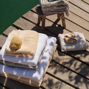 A set of folded Egyptian cotton bath towels and bath mats in cream, white, green and light grey, arranged on a wooden dock next to a green body of water. One stack includes a beige loofah sponge and another includes a wooden-handled towel brush. | Scooms