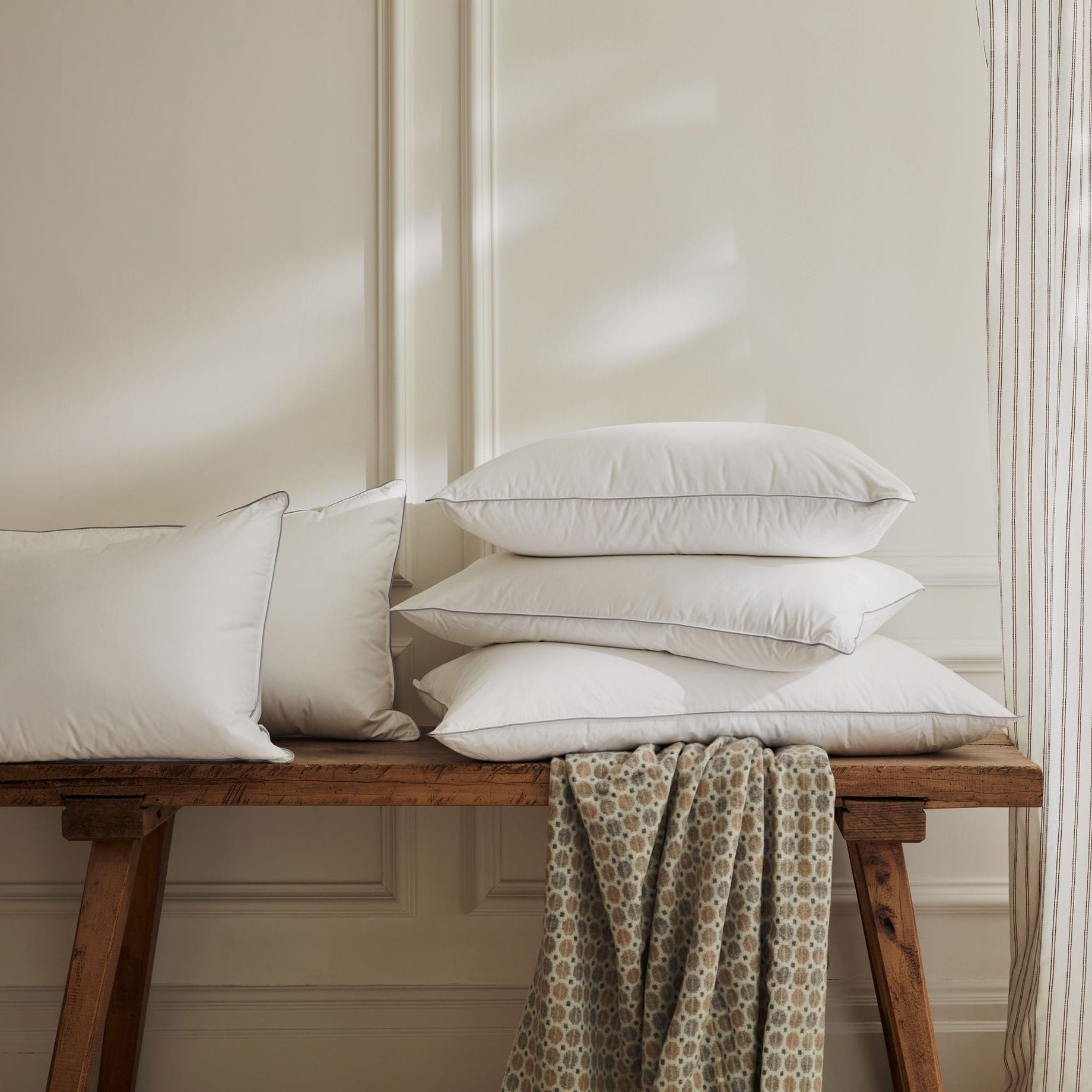 Hungarian goose down pillows on a wooden bench next a window with drapes and a dappled light. Scooms