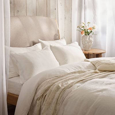 White linen bedding on a bed with linen drapes and a vase of flowers on a side table. | Scooms