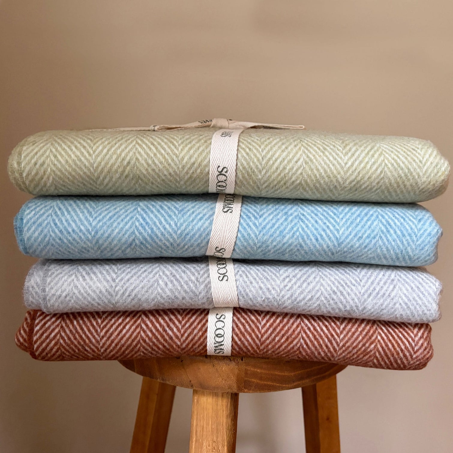 Stack of four folded Merino lambswool throws in different colours on a wooden stool with Scooms branding.