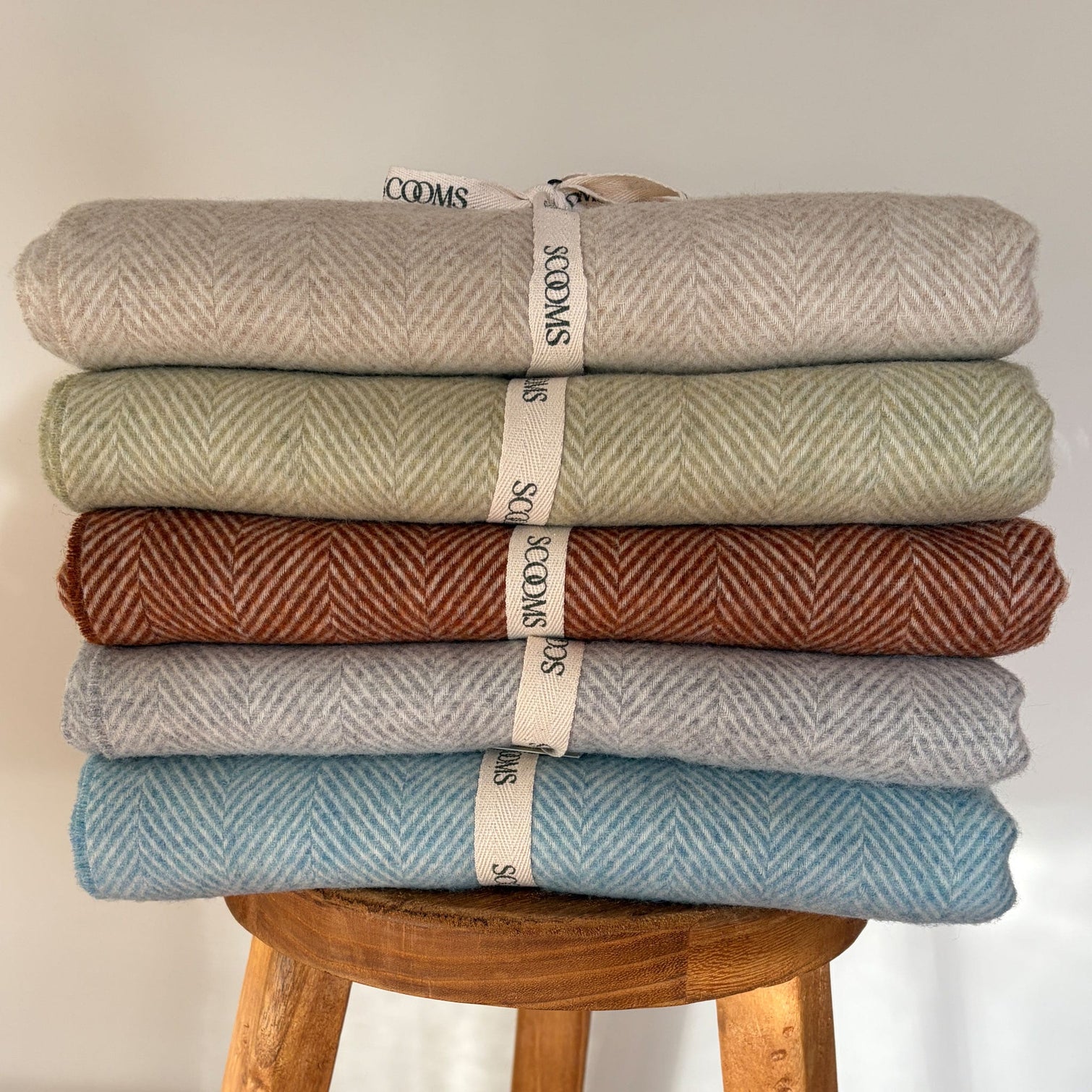 Stack of five folded Merino lambswool throws in different colours on a wooden stool with Scooms branding.