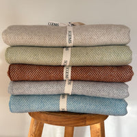 Stack of five folded Merino lambswool throws in different colours on a wooden stool with Scooms branding.
