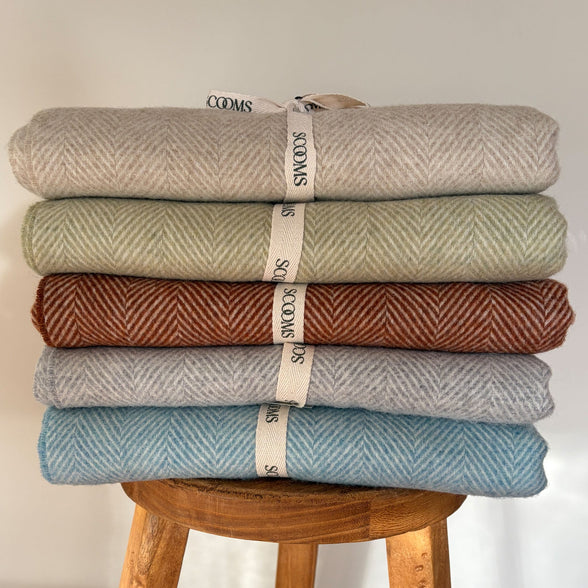 Stack of five folded Merino lambswool throws in different colours on a wooden stool with Scooms branding.