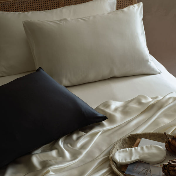 A styled bed scene with ivory Mulberry silk bedding, two ivory pillowcases, and one black pillowcase in the foreground. A matching ivory silk eye mask rests on a woven tray with a book and dried botanicals. | Scooms