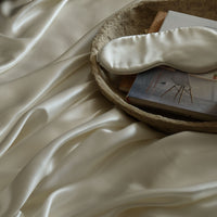 An ivory silk eye mask resting on a tray, on top of a draped ivory Mulberry silk sheet. | Scooms