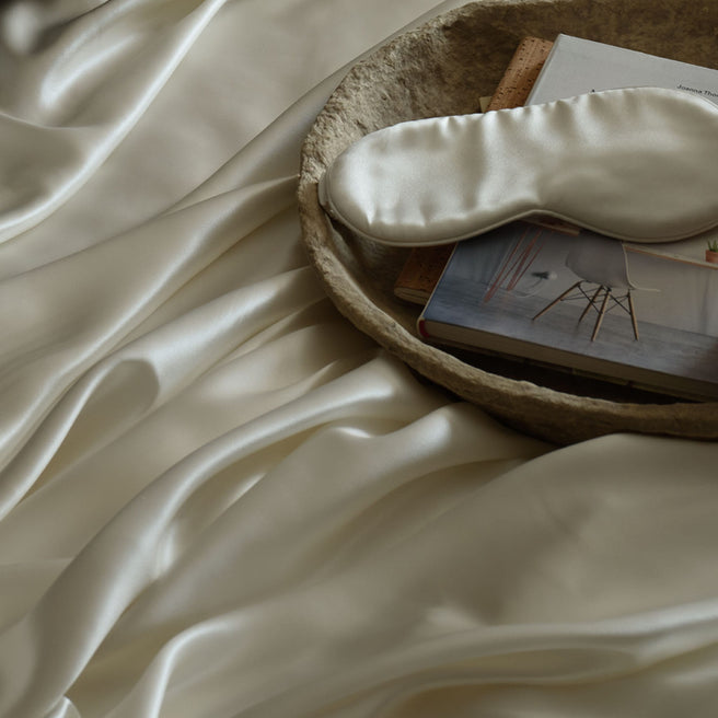 An ivory silk eye mask resting on a tray, on top of a draped ivory Mulberry silk sheet. | Scooms