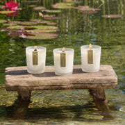 Three natural scented candles in white frosted glass jars sit on a rustic wooden bench over a pond with lily pads and water lilies. | Scooms