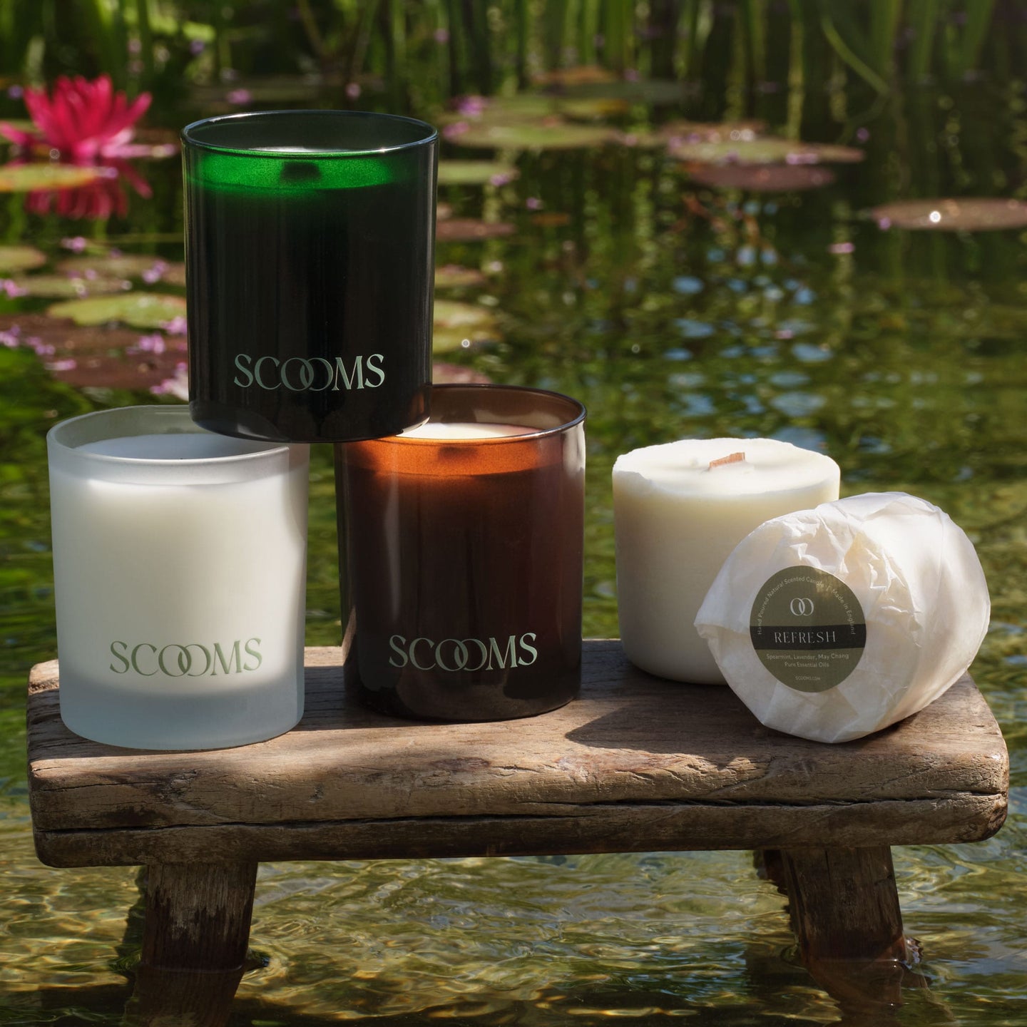 Three Scooms scented candles, plus two candle refills, arranged on a rustic wooden bench over shallow water with lily pads and reeds in the background. The candles feature frosted white, dark amber and green glass jars. | Scooms