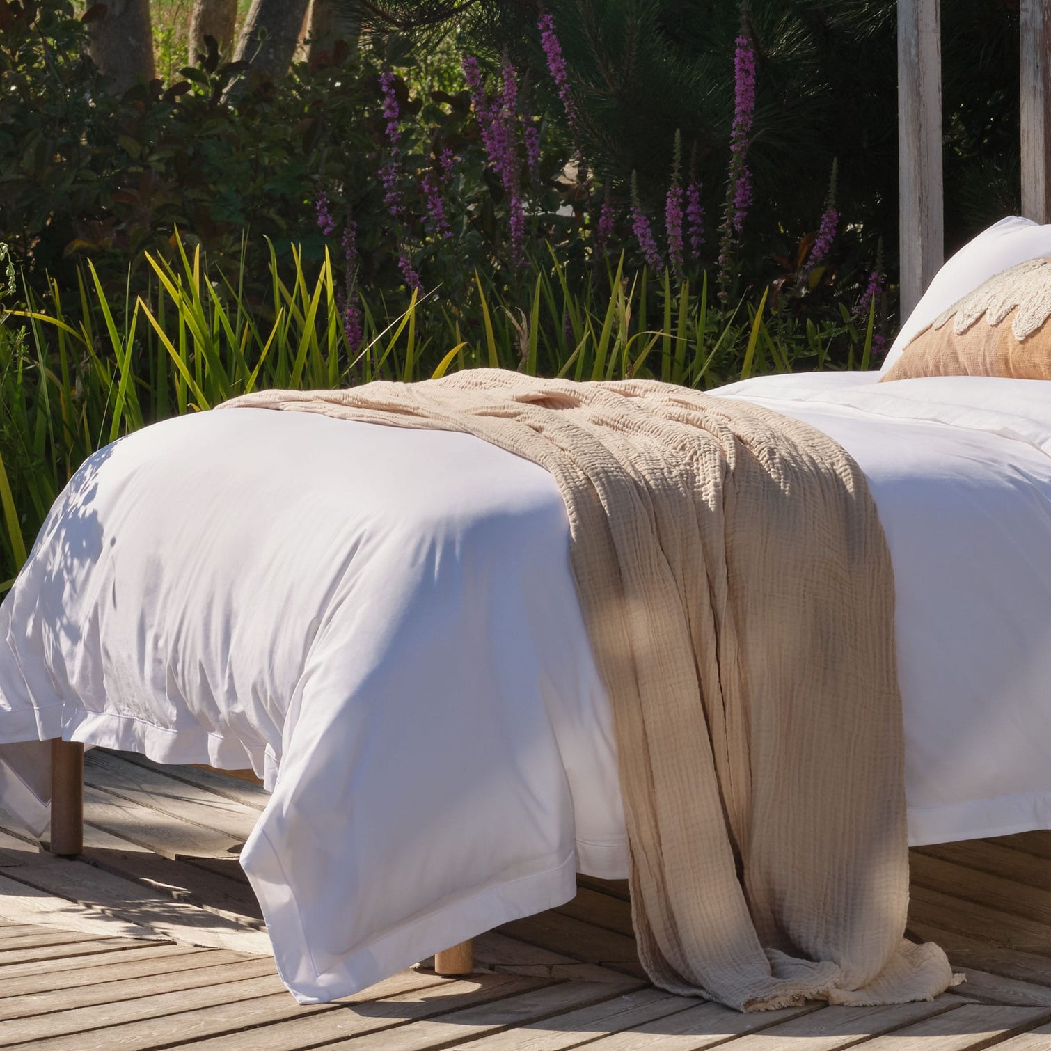 A white bed with Scooms crisp percale cotton sheets and a beige textured throw blanket draped across it, placed on a wooden deck with green plants and purple flowers in the background | Scooms
