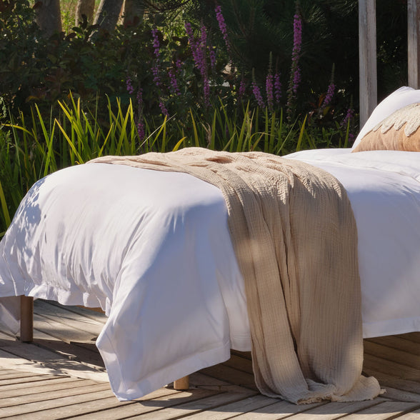 A white bed with Scooms crisp percale cotton sheets and a beige textured throw blanket draped across it, placed on a wooden deck with green plants and purple flowers in the background | Scooms