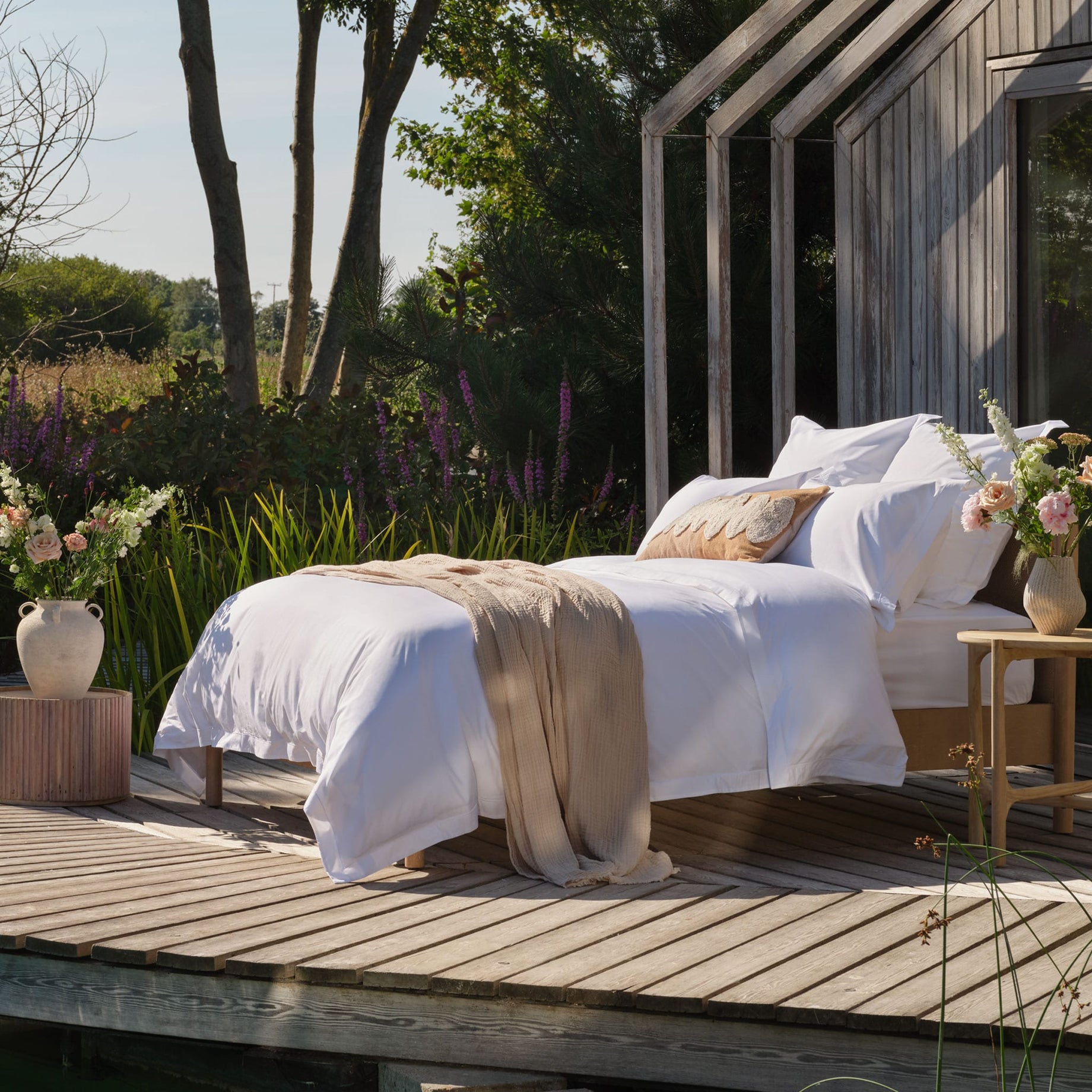 A neatly made bed with white percale bedding and a natural throw blanket sits on a wooden deck beside a modern wooden house, with flowers, greenery, and trees in the background | Scooms