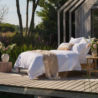 A neatly made bed with white percale bedding and a natural throw blanket sits on a wooden deck beside a modern wooden house, with flowers, greenery, and trees in the background | Scooms