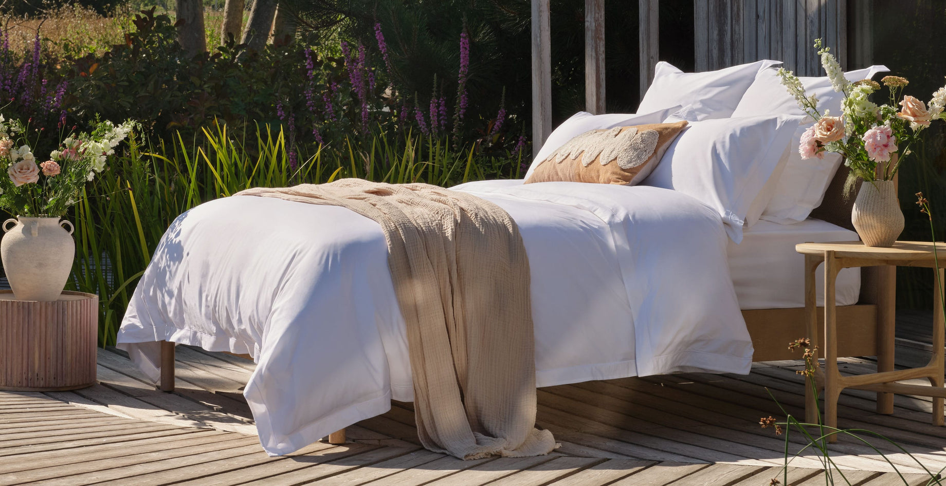A luxurious neatly made bed with white percale Egyptian cotton bedding and a beige throw blanket sits on a wooden deck outdoors, with flowers, greenery, and a wooden structure in the background |Scooms
