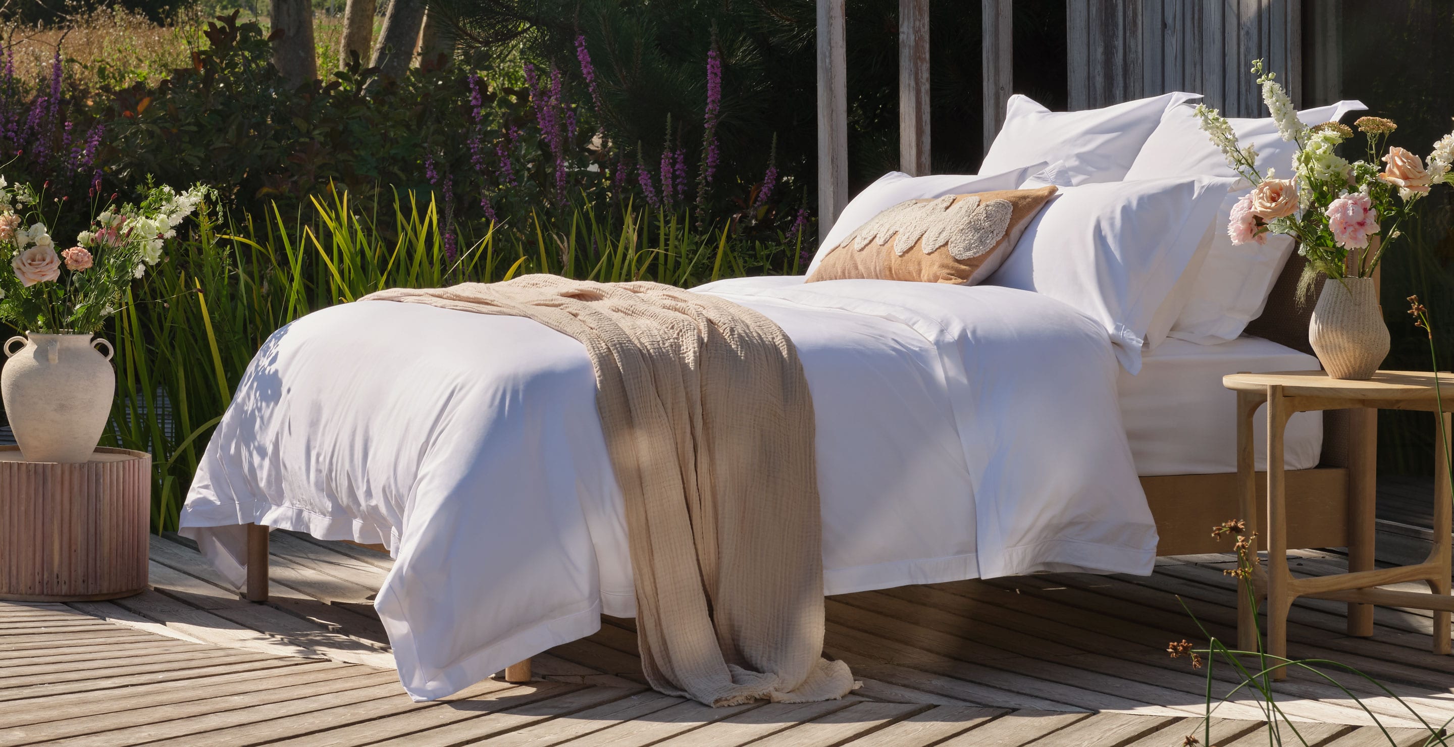 A luxurious neatly made bed with white percale Egyptian cotton bedding and a beige throw blanket sits on a wooden deck outdoors, with flowers, greenery, and a wooden structure in the background |Scooms
