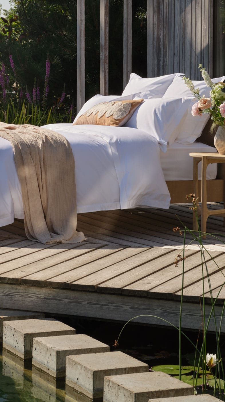 A luxury outdoor bed setup on a wooden deck next to a pool, featuring percale Egyptian cotton white bedding, beige accents, and a modern wooden cabin in a lush green setting. Scooms