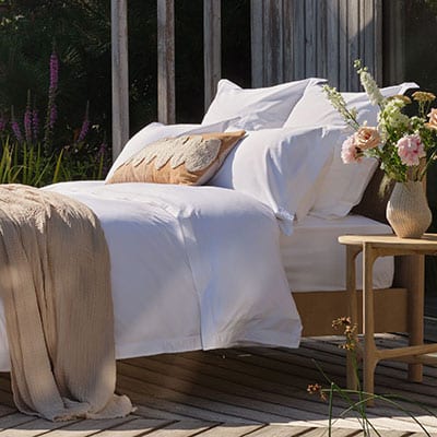 A neatly made bed with white percale bedding and pillows, including a decorative beige pillow, set on a wooden deck beside a small table with a vase of pink flowers. A beige throw blanket is draped over the foot of the bed, with a rustic wooden fence and garden foliage in the background. | Scooms