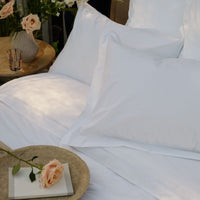 White percale Egyptian cotton bed linens with pillowcases and a white sheet, styled with a woven tray holding a book and a peach rose, with a natural textured throw in the foreground | Scooms