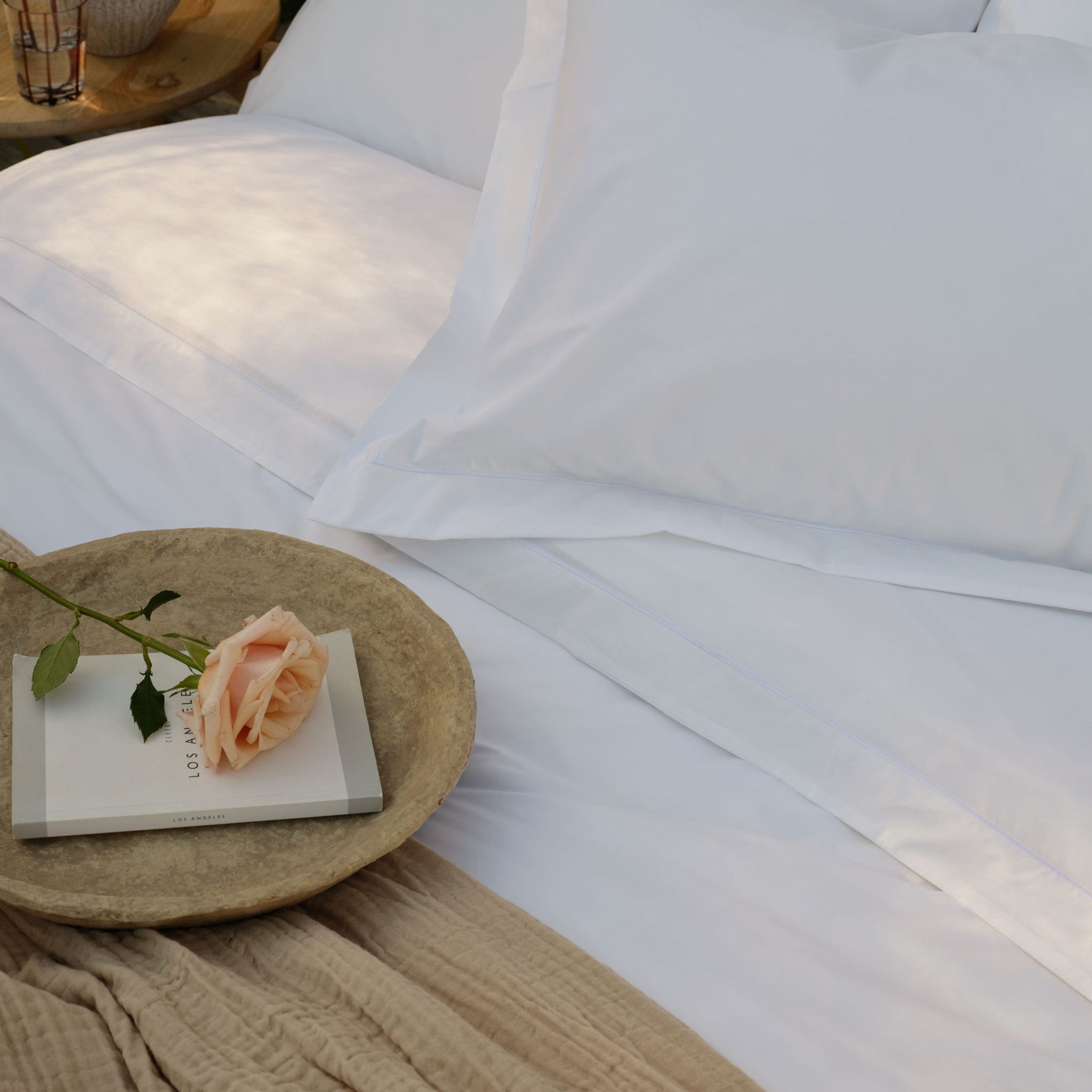 White percale Egyptian cotton bed linens with pillowcases and a white sheet, styled with a woven tray holding a book and a peach rose, with a natural textured throw in the foreground | Scooms
